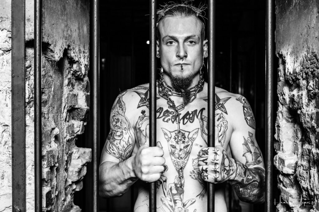 A tattooed man is holding the bars of a cell door at SPNKD.